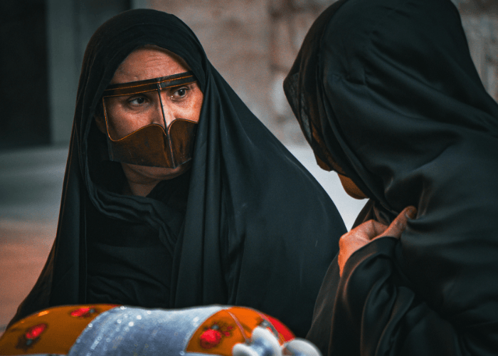 Two Omani women in black abayas and traditional gold burqa face masks in quiet conversation at a market