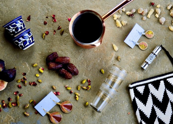 Flatlay of Pink Jinn Souq products including a copper coffee pot, dates, blue ceramic cups, gold earrings, a Moroccan tea glass and a woven clutch bag